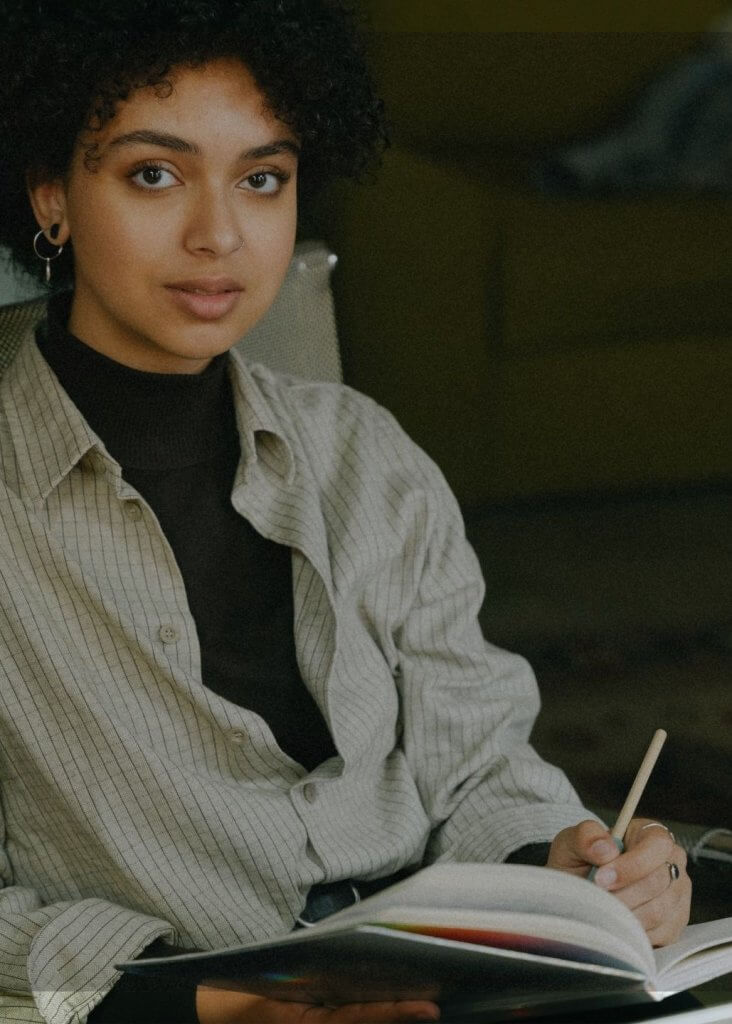 Personne assise, regarde l&rsquo;objectif, &eacute;crit dans un carnet ouvert avec un stylo.